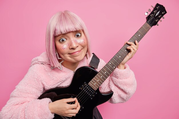 Heureux femme aux cheveux roses joue de la guitare électrique exécute le genre de musique préféré a le visage décoré de paillettes vêtus d'un manteau