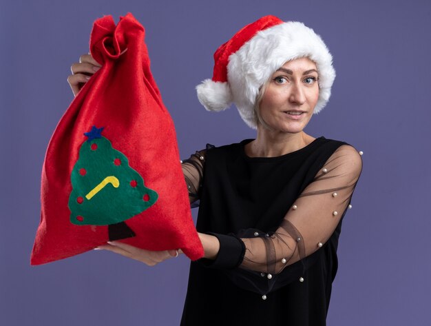 Heureux femme blonde d'âge moyen portant un chapeau de Noël étendant le sac de Noël vers la caméra regardant la caméra isolée sur fond violet