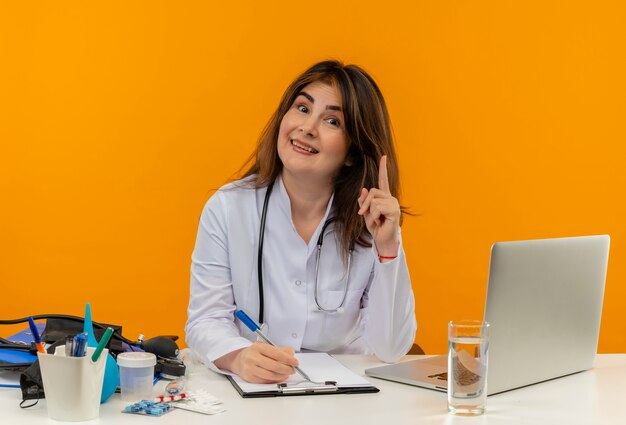 Heureux femme médecin d'âge moyen portant une robe médicale avec stéthoscope assis au bureau de travail sur un ordinateur portable avec des outils médicaux écrire sur des points de presse-papiers vers le haut sur un mur orange isolé avec espace de copie