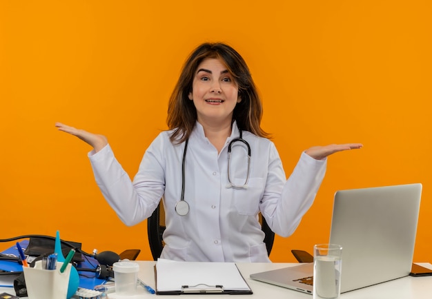 Heureux Femme Médecin D'âge Moyen Portant Une Robe Médicale Avec Stéthoscope Assis Au Bureau De Travail Sur Un Ordinateur Portable Avec Des Outils Médicaux Se Propage Les Mains Sur Un Mur Orange Isolé Avec Espace De Copie