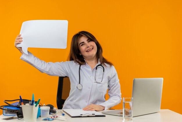 Heureux femme médecin d'âge moyen portant une robe médicale avec stéthoscope assis au bureau de travail sur un ordinateur portable avec des outils médicaux tenant une bulle de discussion sur un mur orange isolé avec espace de copie