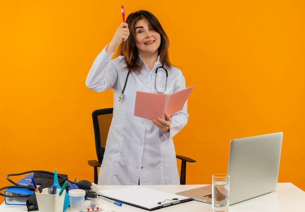 Heureux femme médecin d'âge moyen portant une robe médicale avec stéthoscope assis au bureau de travail sur un ordinateur portable avec des outils médicaux tenant un cahier et un stylo sur un mur orange isolé avec espace de copie