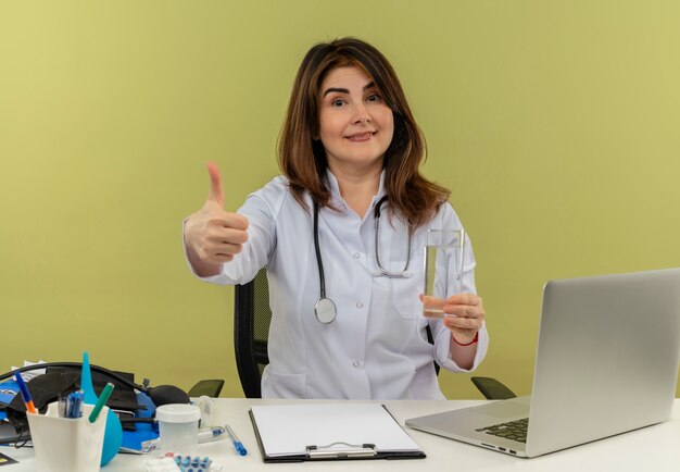 Heureux femme médecin d'âge moyen portant une robe médicale avec stéthoscope assis au bureau de travail sur un ordinateur portable avec des outils médicaux tenant un verre d'eau son pouce vers le haut sur un mur vert avec espace copie