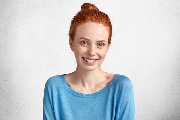 Heureux femme souriante avec une peau tachetée de rousseur et un sourire positif, vêtue d'un pull décontracté bleu, étant en pleine forme après une date avec son petit ami, isolé sur blanc