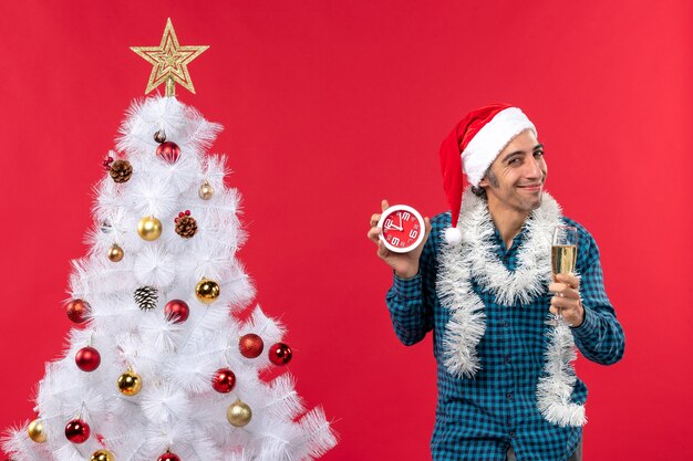 Heureux, fier, jeune homme, à, chapeau père noël, et, lever, a, verre vin, et, tenue, horloge, debout, près, arbre noël, sur, rouge Banque de Photo