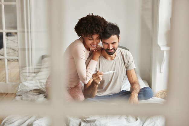 Heureux futurs parents métis regardent joyeusement le test de grossesse, se réjouissent des nouvelles positives de la grossesse, s'assoient ensemble sur le lit contre l'intérieur domestique.