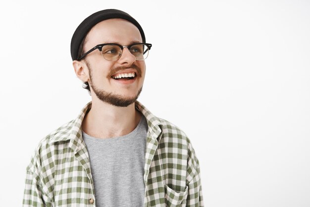 Heureux heureux et ravi garçon patineur masculin chanceux dans des lunettes de bonnet noir et chemise décontractée à carreaux à droite avec un large sourire satisfait écoute compagnon racontant une histoire drôle sur un mur gris