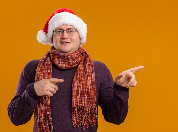 Heureux homme adulte portant des lunettes et bonnet de Noel avec foulard autour du cou regardant la caméra pointant sur le côté isolé sur fond orange