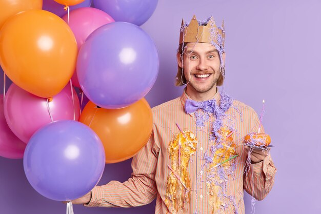 Heureux homme adulte sale avec de la crème de gâteau tient un petit gâteau avec une bougie s'amuse à l'enterrement de vie de garçon porte une couronne de papier sur la tête tient des ballons gonflés colorés isolés sur un mur violet