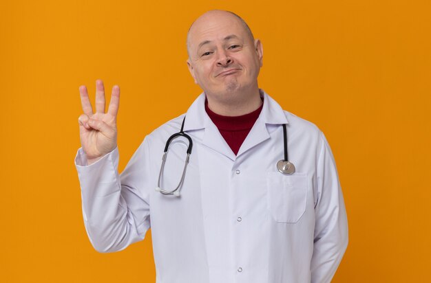 Heureux homme adulte en uniforme de médecin avec stéthoscope gesticulant trois avec ses doigts