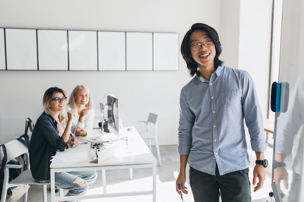 Heureux homme d'affaires asiatique regardant flipchart avec sourire debout dans la salle de conférence. Charmantes étudiantes blondes avec des ordinateurs portables regardant un jeune enseignant écrire à bord.