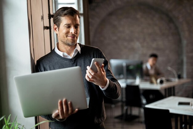 Heureux homme d'affaires debout près de la fenêtre du bureau et lisant un message texte sur un téléphone portable tout en tenant un ordinateur portable