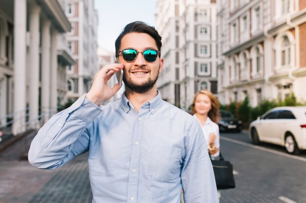 Heureux homme d'affaires à lunettes de soleil parlant au téléphone dans la rue. Jolie fille blonde l'attrapant par derrière