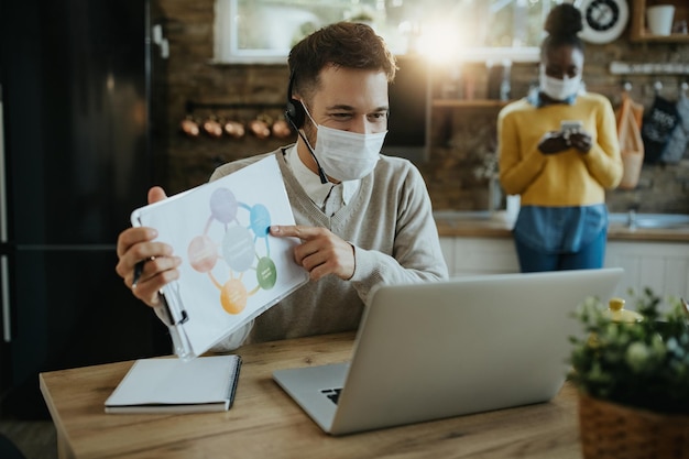 Heureux homme d'affaires avec masque facial analysant le graphique lors d'un appel vidéo à la maison