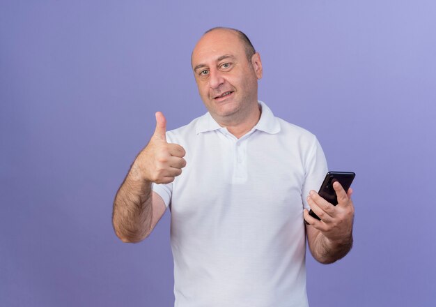 Heureux homme d'affaires mature occasionnel regardant la caméra tenant le téléphone mobile et montrant le pouce vers le haut isolé sur fond violet avec espace de copie