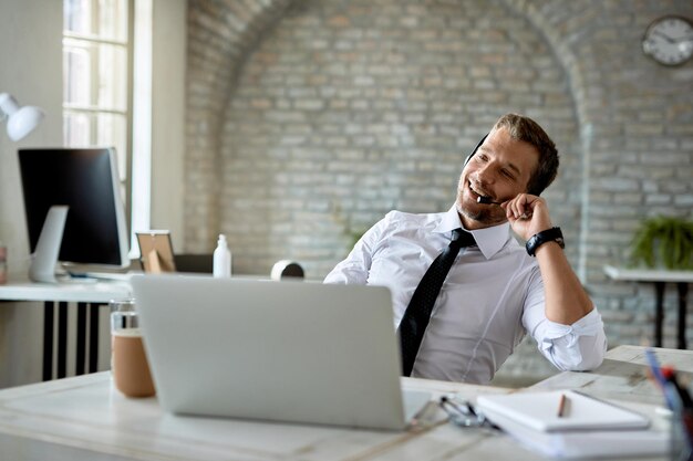 Heureux homme d'affaires portant un casque tout en communiquant avec quelqu'un et en utilisant un ordinateur au bureau