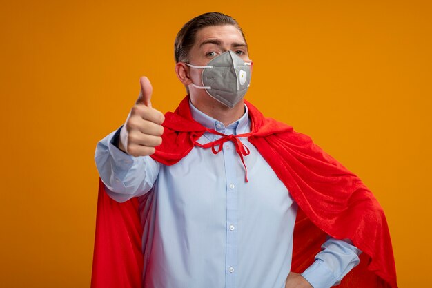 Heureux homme d'affaires de super héros en masque facial protecteur et cape rouge regardant la caméra montrant les pouces vers le haut debout sur fond orange