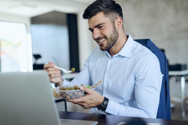 Heureux homme d'affaires surfant sur le net sur un ordinateur portable tout en mangeant des aliments sains pendant la pause déjeuner