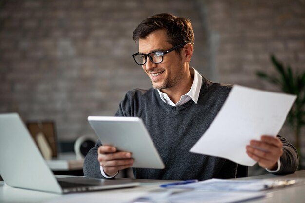 Heureux homme d'affaires utilisant un pavé tactile et un ordinateur portable tout en travaillant sur des rapports d'activité au bureau