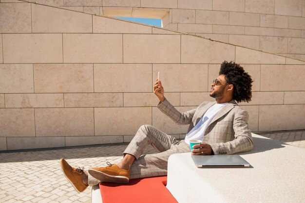 Heureux homme afro-américain appelant en plein air. Homme en costume avec barbe tenant un téléphone portable. Assis en terrasse ou sur les toits. Buvant du café. Communication, concept technologique