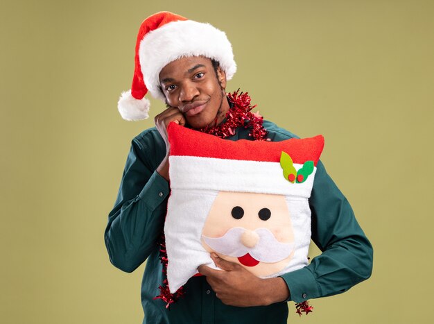 Heureux homme afro-américain en bonnet de noel avec guirlande tenant l'oreiller de Noël regardant la caméra avec sourire sur le visage debout sur fond vert