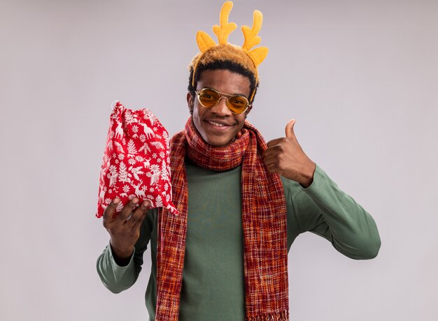 Heureux Homme Afro-américain Avec Jante Drôle Avec Des Cornes De Cerf Et Une écharpe Autour Du Cou Tenant Un Sac De Père Noël Rouge Avec Des Cadeaux Regardant La Caméra En Souriant Montrant Les Pouces Vers Le Haut Debout Sur Fond Vert