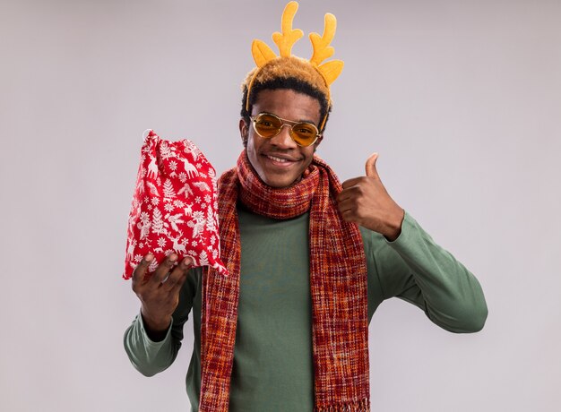 Heureux homme afro-américain avec jante drôle avec des cornes de cerf et une écharpe autour du cou tenant un sac de père Noël rouge avec des cadeaux regardant la caméra en souriant montrant les pouces vers le haut debout sur fond vert