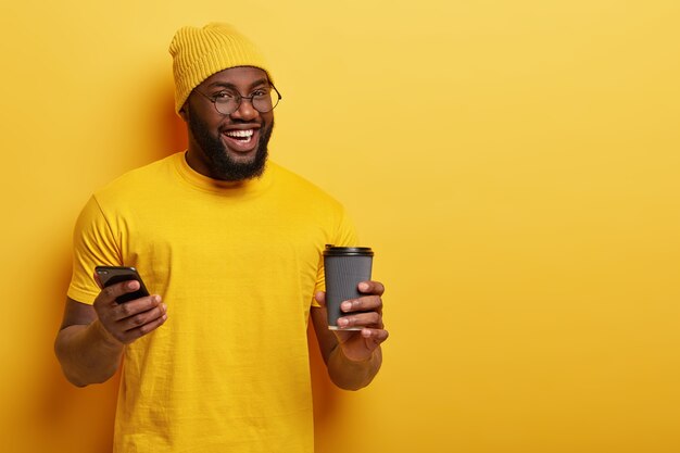 Heureux homme afro-américain lit le fil de nouvelles dans les réseaux sociaux, profite d'une boisson chaude dans un gobelet jetable