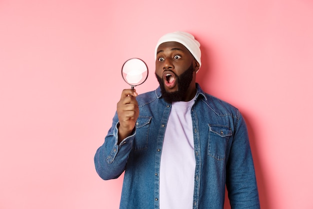 Heureux homme afro-américain regardant à travers une loupe, souriant étonné, debout sur fond rose.