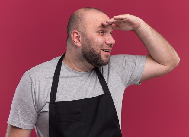 Heureux homme d'âge moyen barbier slave en uniforme à la recherche à distance avec main isolé sur mur rose