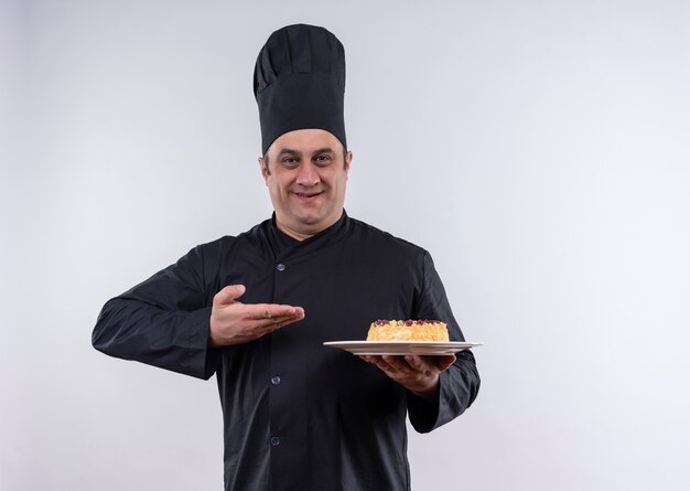 Heureux homme d'âge moyen cuisinier en uniforme de chef montrant un gâteau sur une assiette à la main sur un mur blanc isolé avec copie espace