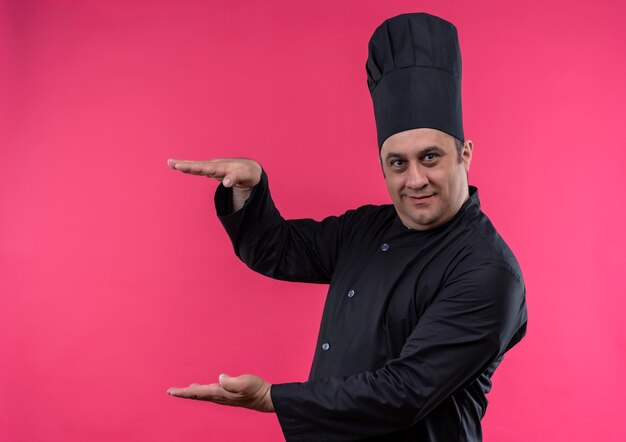 Heureux homme d'âge moyen cuisinier en uniforme de chef montrant la taille sur un mur rose isolé avec copie espace