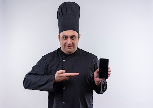 Heureux homme d'âge moyen cuisinier en uniforme de chef montrant le téléphone dans sa main sur un mur blanc isolé