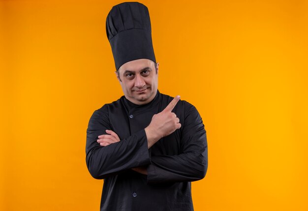 Heureux homme d'âge moyen cuisinier en uniforme de chef pointe doigt à côté sur le mur jaune avec copie espace