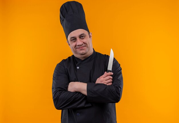 Heureux homme d'âge moyen cuisinier en uniforme de chef tenant un couteau croisant les mains sur le mur jaune