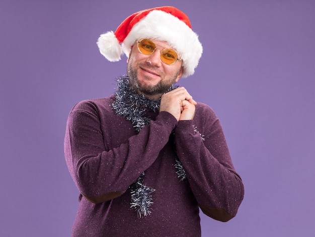 Heureux homme d'âge moyen portant bonnet de Noel et guirlande de guirlandes autour du cou avec des lunettes gardant les mains ensemble isolé sur mur violet