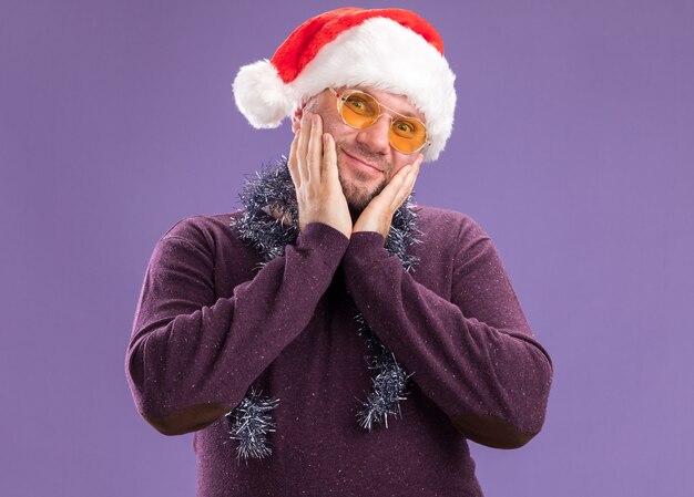 Heureux homme d'âge moyen portant bonnet de Noel et guirlande de guirlandes autour du cou avec des lunettes en gardant les mains sur le visage en regardant la caméra isolée sur fond violet