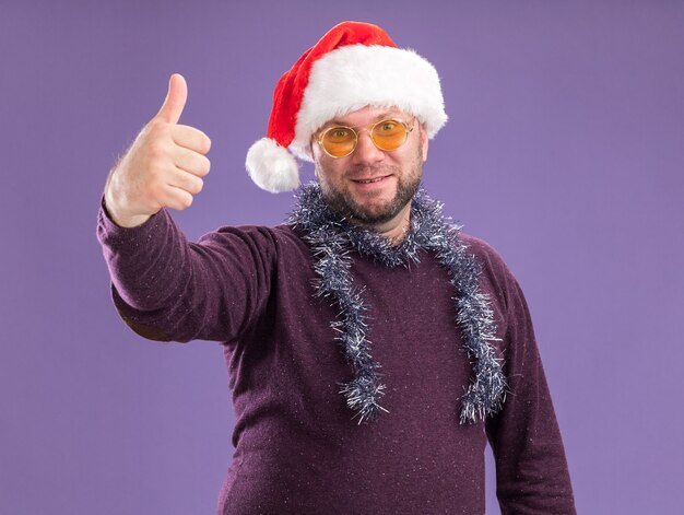 Heureux homme d'âge moyen portant bonnet de Noel et guirlande de guirlandes autour du cou avec des lunettes montrant le pouce vers le haut isolé sur mur violet