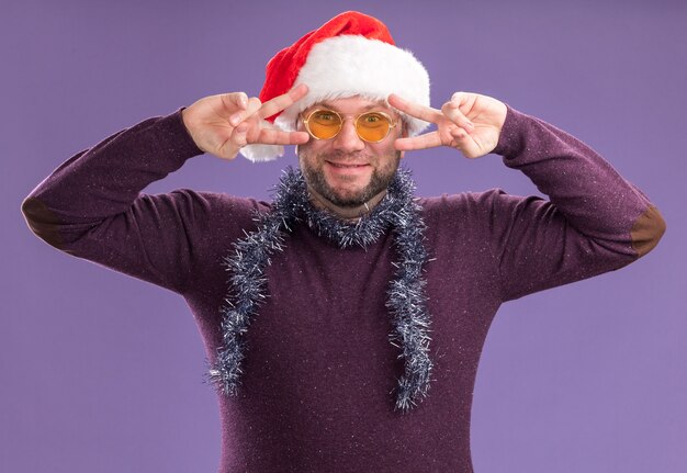 Heureux homme d'âge moyen portant bonnet de Noel et guirlande de guirlandes autour du cou avec des lunettes regardant la caméra montrant des symboles v-sign