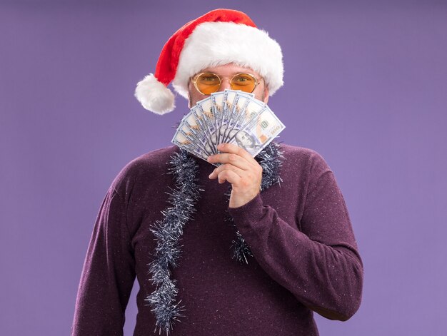 Heureux homme d'âge moyen portant bonnet de Noel et guirlande de guirlandes autour du cou avec des lunettes tenant de l'argent par derrière isolé sur mur violet