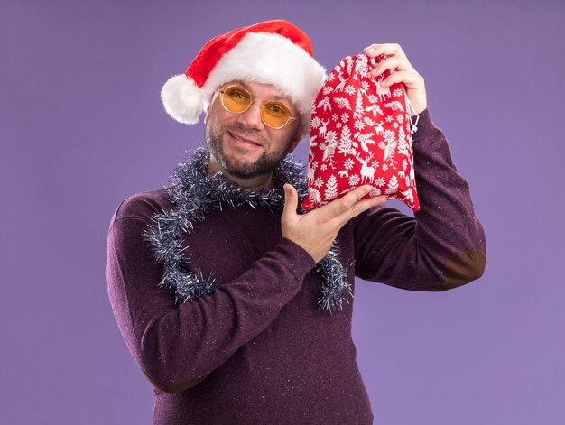 Heureux homme d'âge moyen portant bonnet de Noel et guirlande de guirlandes autour du cou avec des lunettes tenant le sac de cadeau de Noël près de la tête isolée sur le mur violet