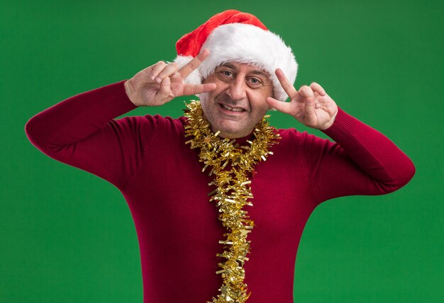 Heureux homme d'âge moyen portant un bonnet de noel de noël avec des guirlandes autour du cou souriant montrant un signe v près des yeux debout sur un mur vert