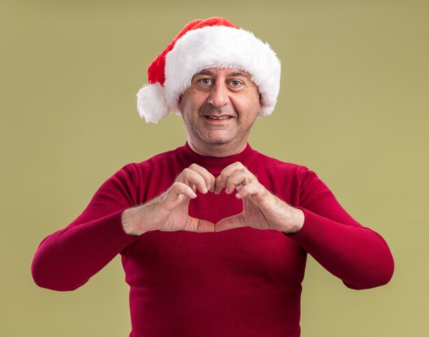 Heureux homme d'âge moyen portant noël bonnet de noel faisant le geste du cœur souriant joyeusement debout sur fond vert