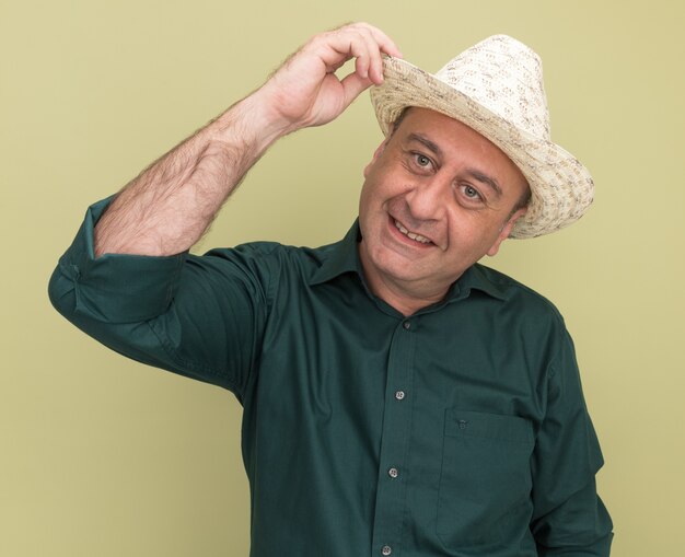 Heureux homme d'âge moyen portant un t-shirt vert et un chapeau isolé sur un mur vert olive
