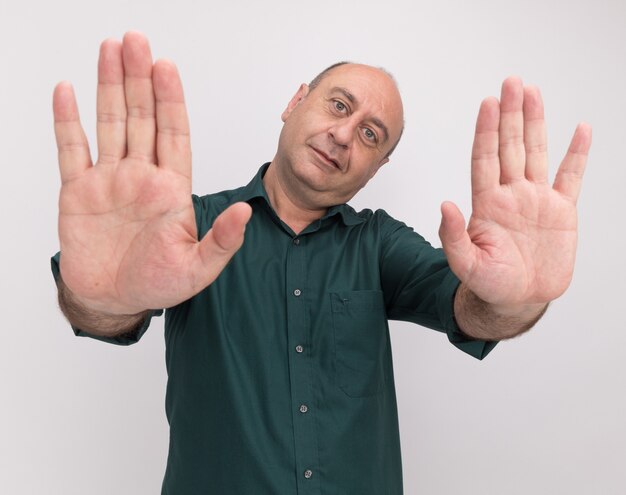 Heureux homme d'âge moyen à tête inclinable portant un t-shirt vert montrant un geste d'arrêt isolé sur un mur blanc