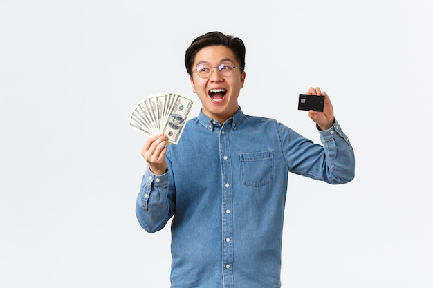 Heureux homme asiatique souriant avec bretelles et lunettes riant joyeusement et montrant une carte de crédit tenant ...