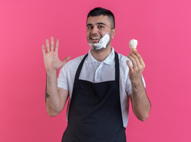 Heureux homme barbier en tablier mettant de la mousse à raser sur son visage avec un blaireau à la confiance debout sur fond rose