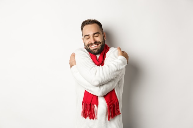 Heureux homme barbu appréciant un pull d'hiver confortable, souriant avec chaleur, se serrant dans ses bras, debout sur fond blanc
