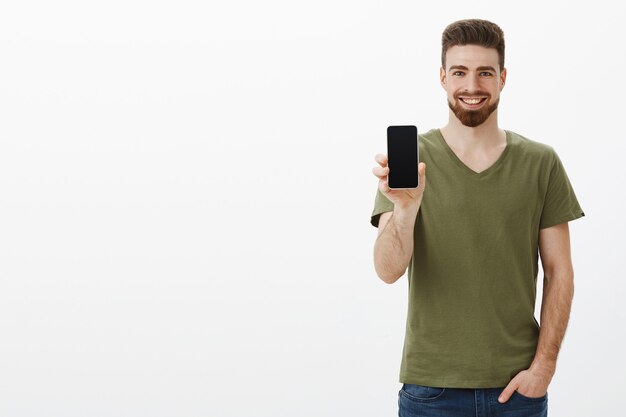 Heureux homme barbu charismatique et confiant, montrant l'écran du smartphone et souriant joyeusement