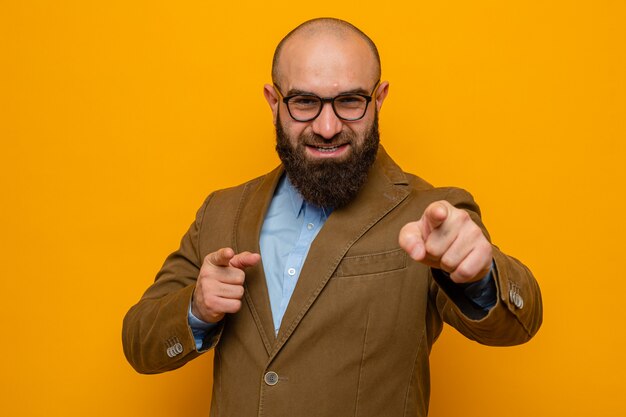 Heureux homme barbu en costume marron portant des lunettes à sourire pointant gaiement avec l'index à l'avant